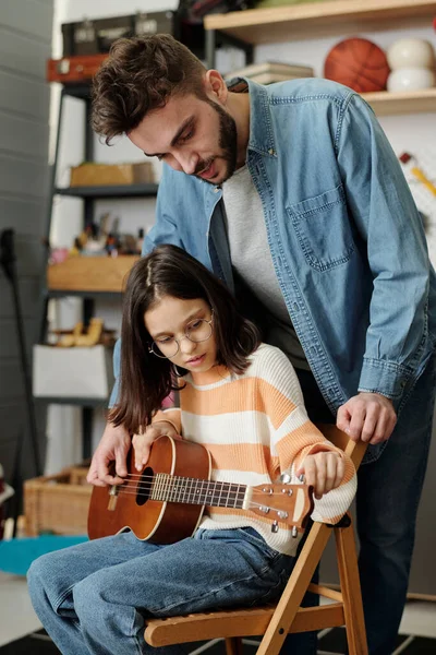 Cute little girl fitting strings of guitar before the lesson of music with her tutor or father in garage while sitting in front of him