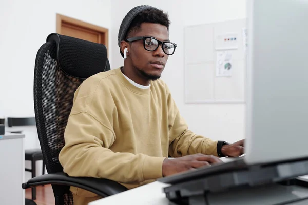 年轻而严肃的男性 穿着黄色运动衫的程序员一边在办公室里解码数据一边看着电脑屏幕 — 图库照片