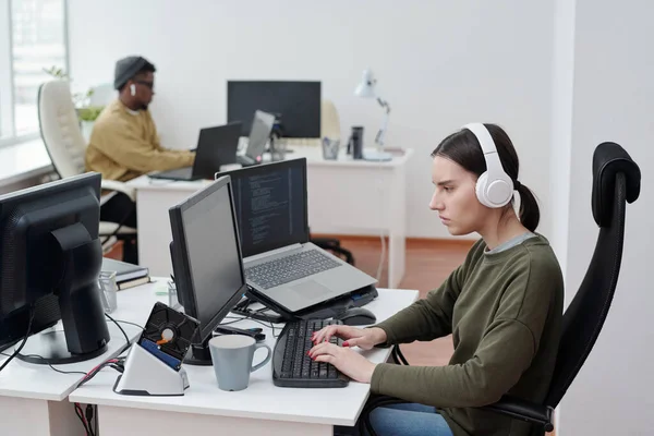 从侧面看耳机里的年轻女程序员在工作场所听音乐时对着在职的非洲裔美国男性同事 — 图库照片