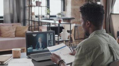 Over-shoulder of young Black man sitting at desk in living room at daytime, video calling mature Caucasian female financier via laptop computer