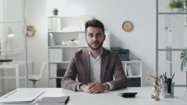 Waist-up of young Caucasian man wearing formal suit, sitting at desk in bright office in afternoon, looking on camera