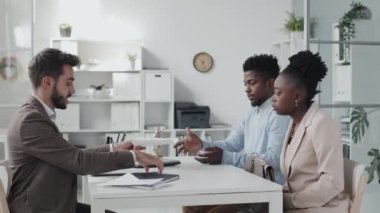 Side view of young African American couple sitting at desk in office, having consultation with male Caucasian lawyer at daytime