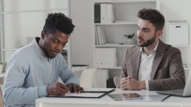 Waist-up of young African American man sitting at desk in office at daytime, signing employment contract with male Caucasian HR manager