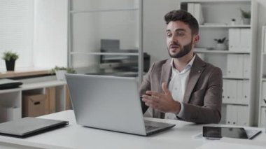 Waist-up of blue-eyed Caucasian entrepreneur sitting at desk in white-colored office in afternoon, video calling via portable computer, using tablet