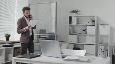 Medium long of blue-eyed Caucasian businessman walking back and forth in white-colored office at daytime, talking on cellphone, holding documents