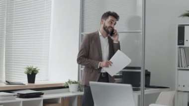 Orta uzunlukta beyaz renkli ofiste yürüyen, cep telefonuyla konuşan, elinde belgelerle gezen Kafkasyalı genç girişimci.