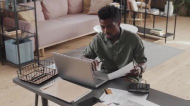 Gündelik giysiler giyen, gündüzleri oturma odasında oturan, bütçe ve vergilerle çalışan, bilgisayar kullanan genç siyahi bir adamın yüksek açılı beline sahip olması.