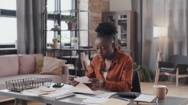 Chest-up of young African American woman opening envelope with invoice, stressing out sitting at desk in living room at daytime
