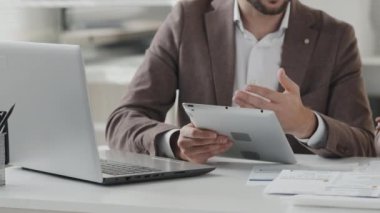 Midsection tracking right of male financial advisor and female client sitting at desk in white office, having business discussion, using tablet computer and documents