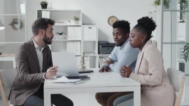 Side-view of blue-eyed Caucasian male counsel and young African American married couple sitting at desk, having business conversation at daytime