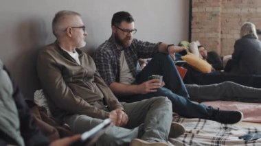 Medium long of two older and younger Caucasian men sitting on mattresses on floor of refuge, having conversation at daytime
