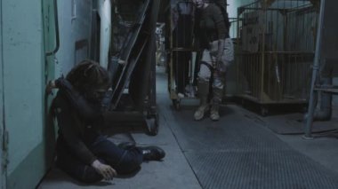 Injured female soldier with shaved head transporting zombies in cage on wheels in dark basement of industrial building