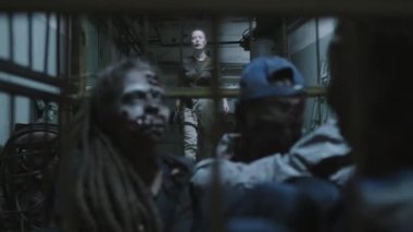 Military woman with shaved head moving away zombies in cage on wheels in dark basement of industrial building