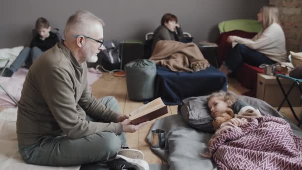 昼間の避難所の床の上にマットレスの上に座っているシニア白人男性の完全なショット 毛布の下に横たわるかわいい孫への本を読んで — ストック動画