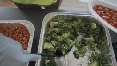 Top view of unrecognizable volunteer in gloves and green reflective vest standing at table indoors at daytime, putting cooked broccoli into disposable lunch box