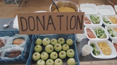 High angle tracking of table with fresh bread, green apples, healthy balanced meals in disposable lunch boxes and handmade cardboard sign with donation inscription indoors at daytime
