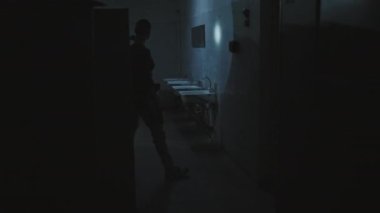 Wide shot of female soldier with shaved head walking in abandoned dark public bathroom with lights flickering, searching first aid kit