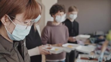 Orta uzunluktaki yüz maskeli mülteciler tek kullanımlık beslenme çantalarını sıcak yemeklerle tutuyorlar, yemek bankasındaki gönüllülerden taze yeşil elma alıyorlar.