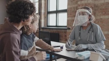 Waist-up of female Biracial nurse in medical gloves and face shield talking to Caucasian woman with little kid sitting at table in indoor shelter at daytime