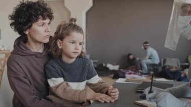 Waist-up of Caucasian woman with little son talking to female Biracial nurse in scrub suit and face shield sitting at table in indoor shelter at daytime