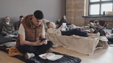 Full shot of dark-haired guy in vest sitting on mat on floor of indoor refugee camp, using smartphone, eating bread