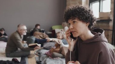 Bulanık mültecilerin öğleden sonra kapalı kampta otururken ön planda telefonla konuşan kıvırcık saçlı beyaz kadının göğüs üstü yan görüntüsü.