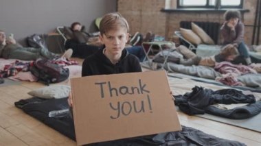 Portrait of blond-haired Caucasian schoolboy holding cardboard sign with thank you inscription on it, looking on camera, sitting in foreground of blurred refugees in indoor camp in afternoon