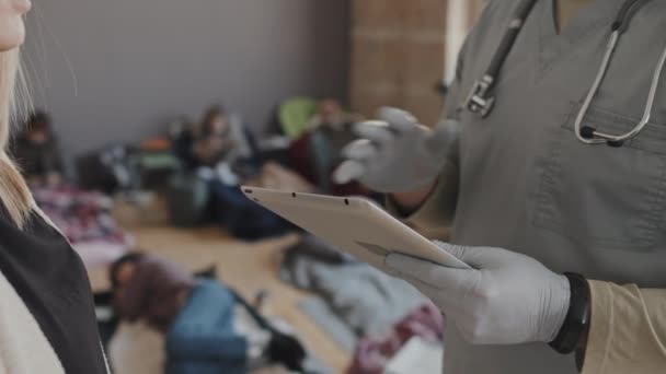 スクラブスーツのクロップド男性医療従事者の中央部とタブレットコンピュータを保持手袋 昼間に亡命中の女性難民と話を — ストック動画