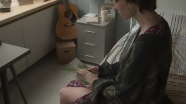 Tilting up of young sad Caucasian female adult with nasal cannula sitting on bed in decorated hospital ward, looking at drawing in her hands