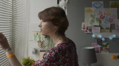 Side view of short-haired Caucasian girl with nasal cannula and dripper looking out window in decorated hospital room