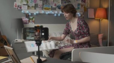 Selective focus of short-haired Caucasian girl with nasal cannula sitting on bed in decorated hospital ward, smiling, talking, showing pill jars, filming vlog on smartphone on tripod