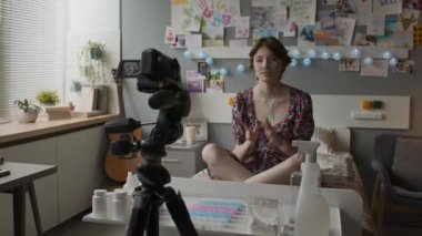 Wide shot of brown-haired Caucasian girl with nasal cannula sitting on bed in decorated hospital ward, smiling, talking, filming vlog on video camera