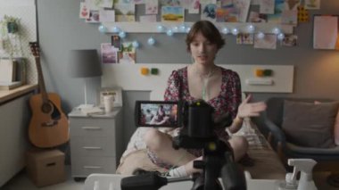Teenage Caucasian girl with nasal cannula sitting on bed in decorated hospital ward, smiling, talking, filming storytime video for online blog on video camera