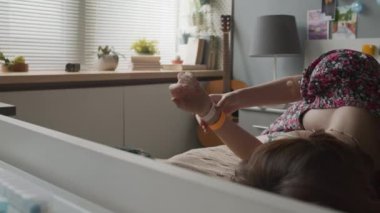 Brown-haired girl lying on bed in ward at daytime, looking at hospital tag on her hand