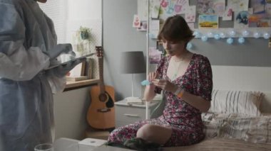 Medium of teenage Caucasian girl sitting on bed in hospital ward in morning, getting pill and glass of water from cropped medical worker standing