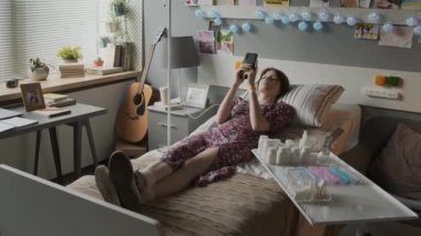 Long shot of bored Caucasian girl wearing dress with floral print and nasal cannula, lying on bed in hospital ward, using smartphone, texting, putting gadget away, looking at pill jars