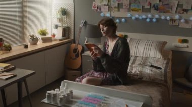 Medium long of short-haired Caucasian girl with nasal cannula sitting on bed in hospital ward in afternoon, reading book and visualizing