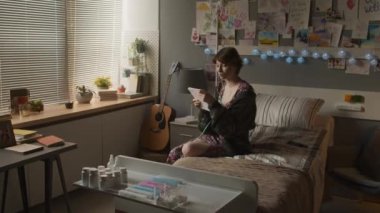 Medium long of teenage girl with nasal cannula and chronic disease sitting on bed in hospital ward in afternoon, taking notes in notebook