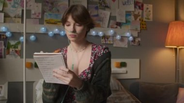 Over-shoulder of short-haired teenage girl with nasal cannula and chronic disease sitting in hospital ward in afternoon, writing in notebook