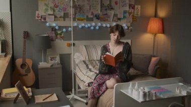 Tracking in portrait of Caucasian girl with nasal cannula and chronic disease sitting on bed in hospital ward in afternoon, reading book, then looking on camera