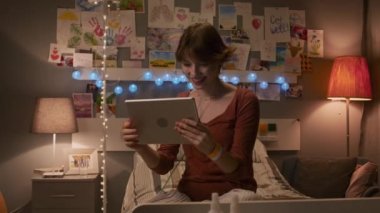 Medium of brown-haired Caucasian young adult with nasal cannula sitting on bed in hospital ward in evening, using tablet computer, video calling, smiling