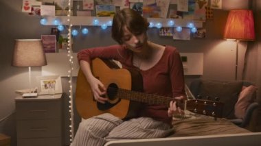 Medium long portrait of Caucasian girl with nasal cannula sitting on bed in hospital room decorated with garlands at night, playing acoustic guitar, then looking up and smiling on camera