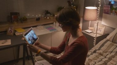 High-angle of Caucasian teenage girl with nasal cannula sitting on bed in hospital ward in evening, using tablet computer, video calling friends, smiling