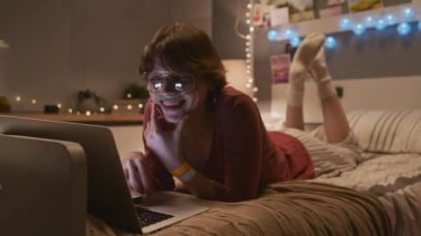 Full shot of young Caucasian woman with nasal cannula lying on bed in decorated hospital room, watching funny online videos on portable computer, laughing