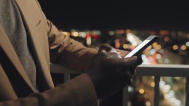 Close-up tilting shot of young black employee standing on balcony late in the evening with blurred cityscape in background and scrolling through messages on corporate smartphone