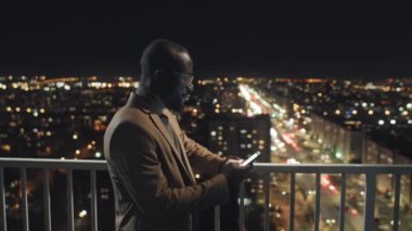 Medium side shot of thoughtful young black office worker in jacket and glasses standing on balcony late at night with city lights in dark blurred background, composing business email on smartphone