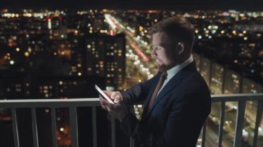 Waist-up shot of young Caucasian male corporate employee dressed in business suit and tie standing on balcony of high office building at night, checking personal messages on smartphone and laughing