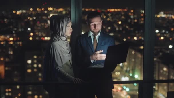 スーツ姿の男とイスラムヒジャーブ姿の女の中出し夜遅く暗い都市部のオフィスで窓のそばに立ってノートパソコンを持ち活発な議論に従事し — ストック動画