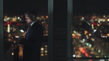 Medium shot of exhausted young businessman in suit standing by office window at night, typing message on smartphone, then rubbing forehead and face. Reflection on glass, bokeh cityscape in background