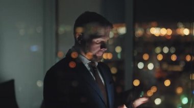 Medium tilting down shot of businessman in suit standing in corporate office late at night, typing on smartphone, rubbing forehead, shaking head, with bokeh lights and female colleague in background